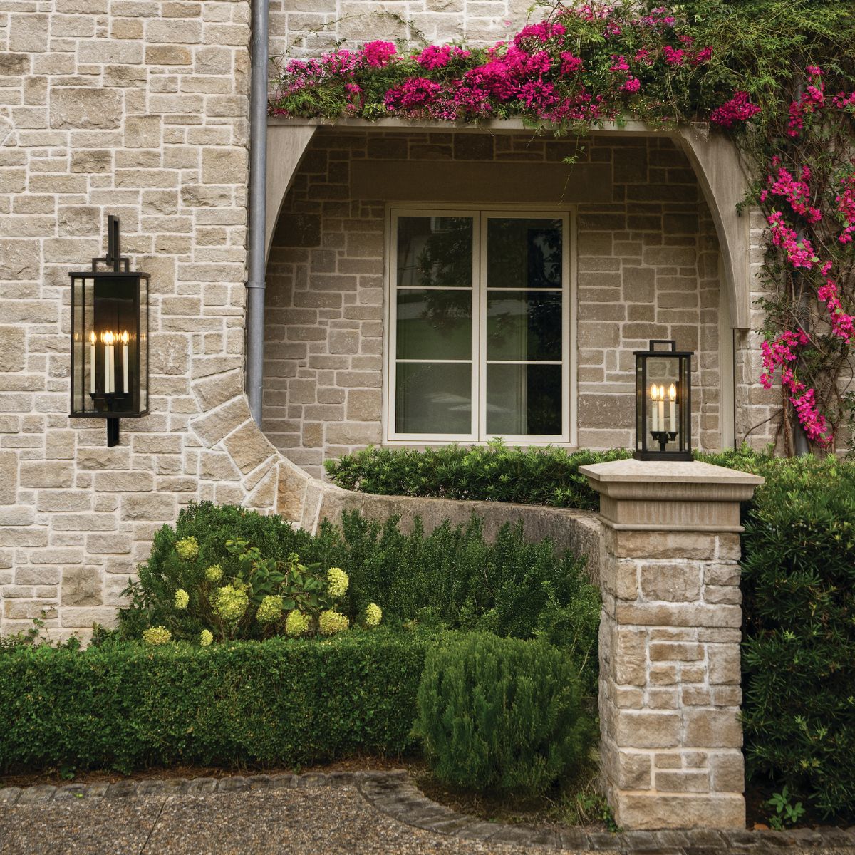 A rustic stone house exterior featuring two black rectangular outdoor wall lanterns, a stone archway, and vibrant pink bougainvillea flowers.